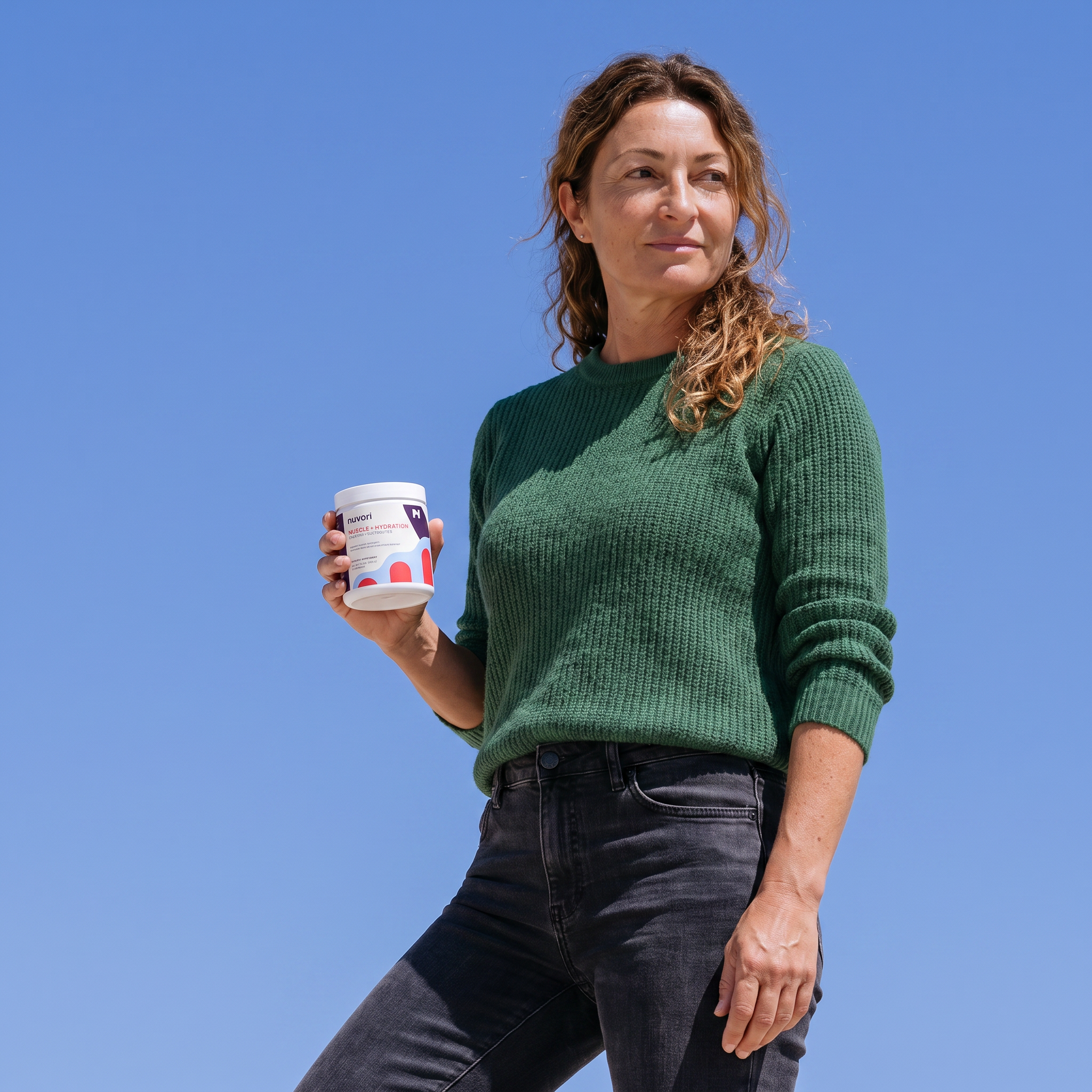 A woman wearing a green ribbed knit sweater and dark jeans stands outdoors against a clear blue sky, holding a white supplement jar with red and black text.